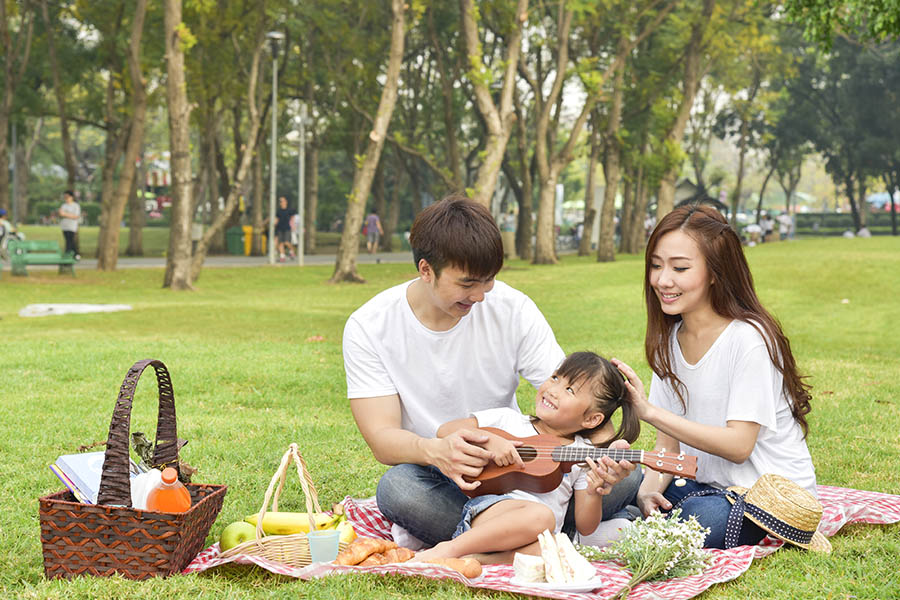 About Our Agency - A Happy Father and Mother are Smiling at Their Daughter Playing With a Small Guitar While Having an Outdoor Picnic at a Park