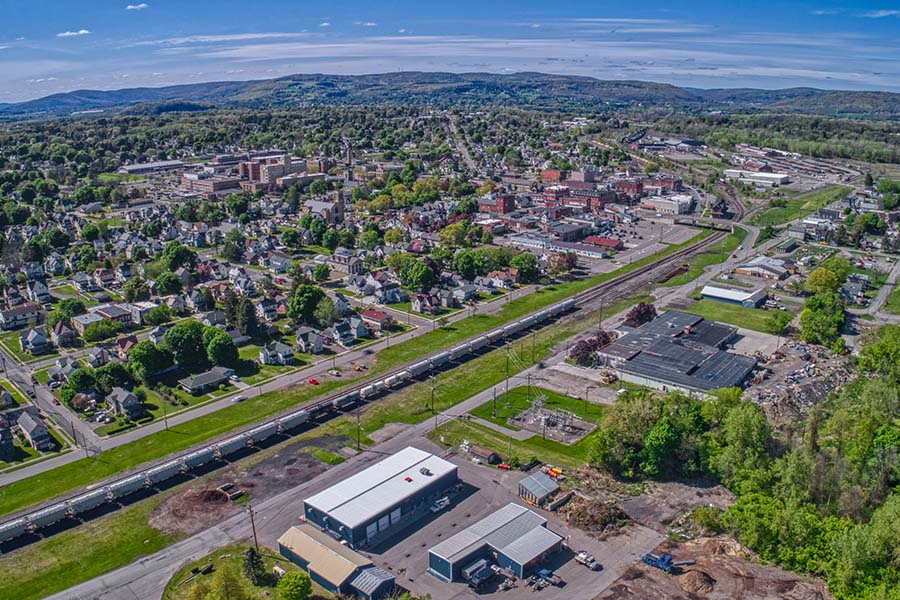 Contact - Aerial View of Sayre, PA Displaying Homes, Buildings, Trees and Mountain Ranges in the Backgrounf on a Sunny Day